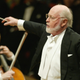 John Williams, dressed in a tuxedo, conducts an orchestra.